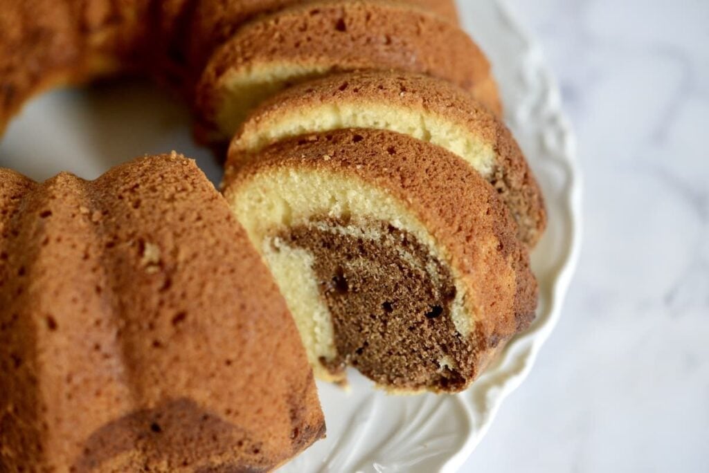 A close-up of a marble bundt cake with several slices cut, showing the swirled pattern of chocolate and vanilla inside, on a white plate with a decorative edge.