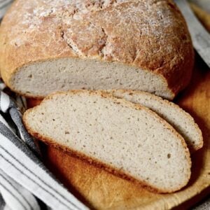 A round loaf of homemade bread with a rustic crust sits on a wooden cutting board. Two slices are cut and laid in front, next to a striped cloth and a knife with a wooden handle.