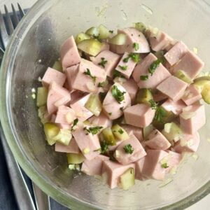 A glass bowl filled with a salad of diced sausage, chopped pickles, onions, and herbs.