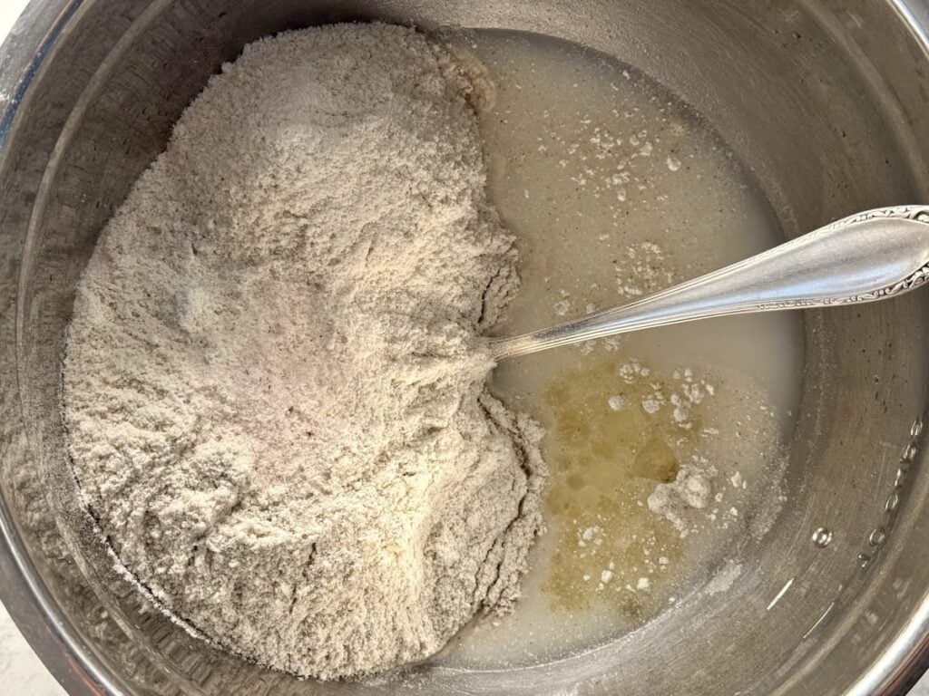A metal bowl containing flour mixture, water, and oil, with a silver spoon partially dipped in the ingredients, ready to be mixed for baking.