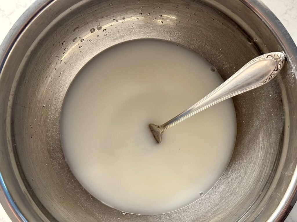 A metal bowl containing a cloudy white liquid with a silver spoon resting inside. The liquid appears to be a mixture, possibly of flour or cornstarch with water.
