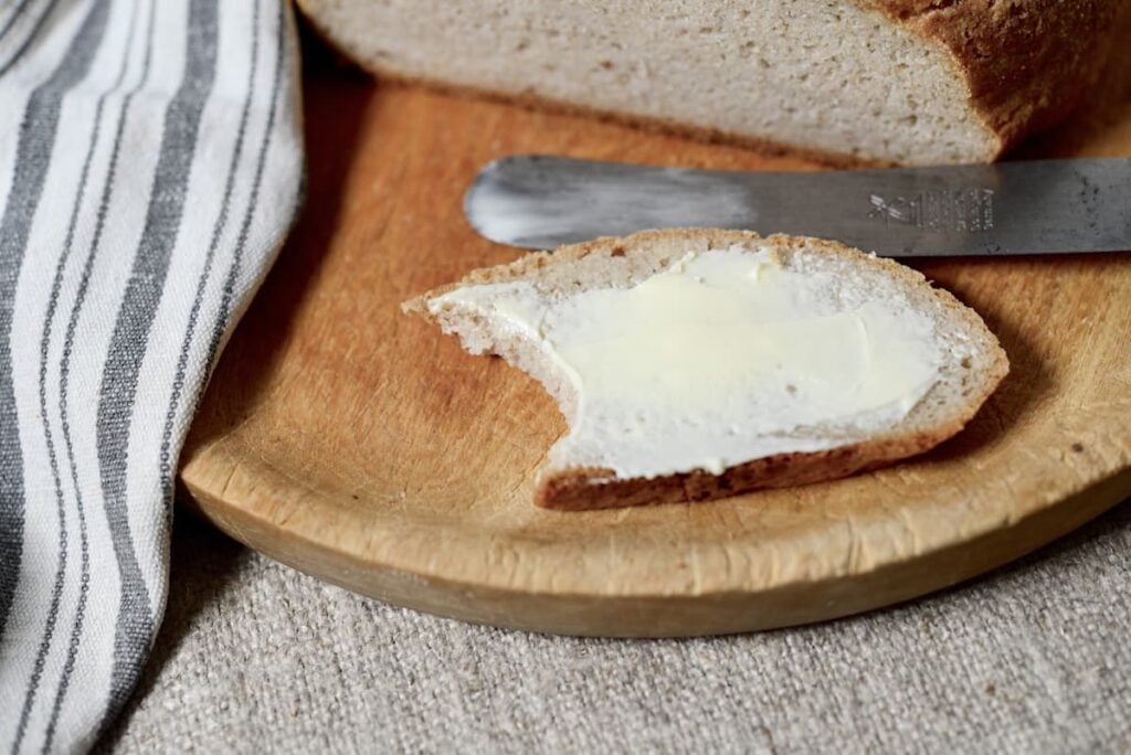 A slice of bread with butter and a bite taken out of it sits on a wooden plate next to a butter knife and a striped cloth napkin.