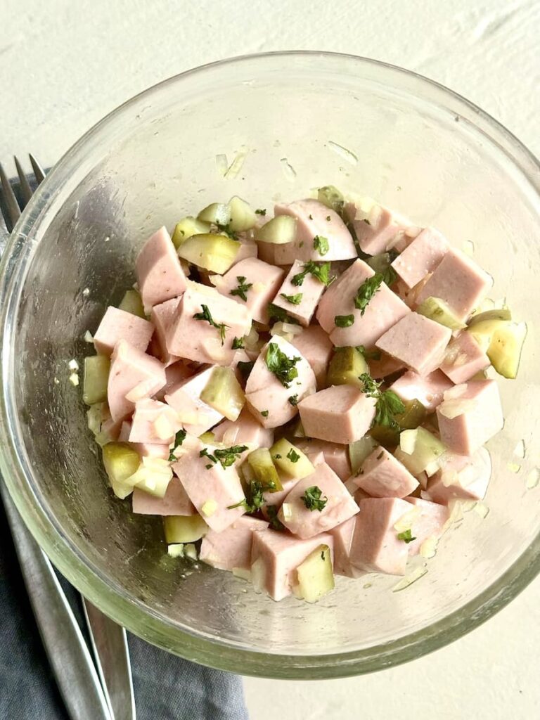 A glass bowl filled with a salad made of diced ham, chopped pickles, and fresh herbs, placed on a light surface next to a gray napkin and silverware.