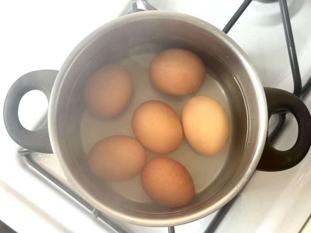 Six brown eggs boiling in a pot of water on a stovetop, with some steam visible on the pot’s surface.