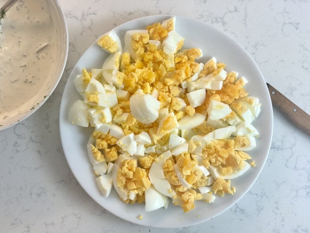 A white plate filled with chopped hard-boiled eggs sits on a light countertop. A bowl of creamy dressing and a knife are nearby.
