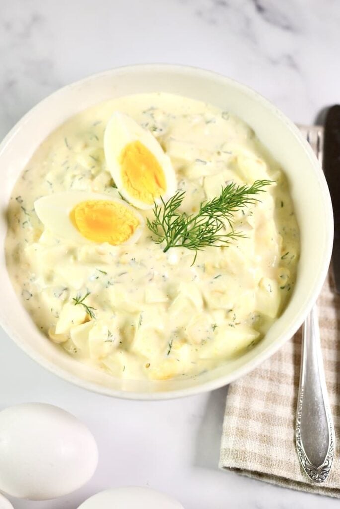 A bowl of creamy egg salad garnished with fresh dill and two egg wedges, placed on a napkin with a spoon beside it. Two whole eggs are visible in the foreground on a white marble surface.