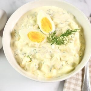 A bowl of creamy egg salad garnished with fresh dill and two egg wedges. Three whole eggs, a knife, and a fork rest nearby on a striped cloth atop a white marble surface.