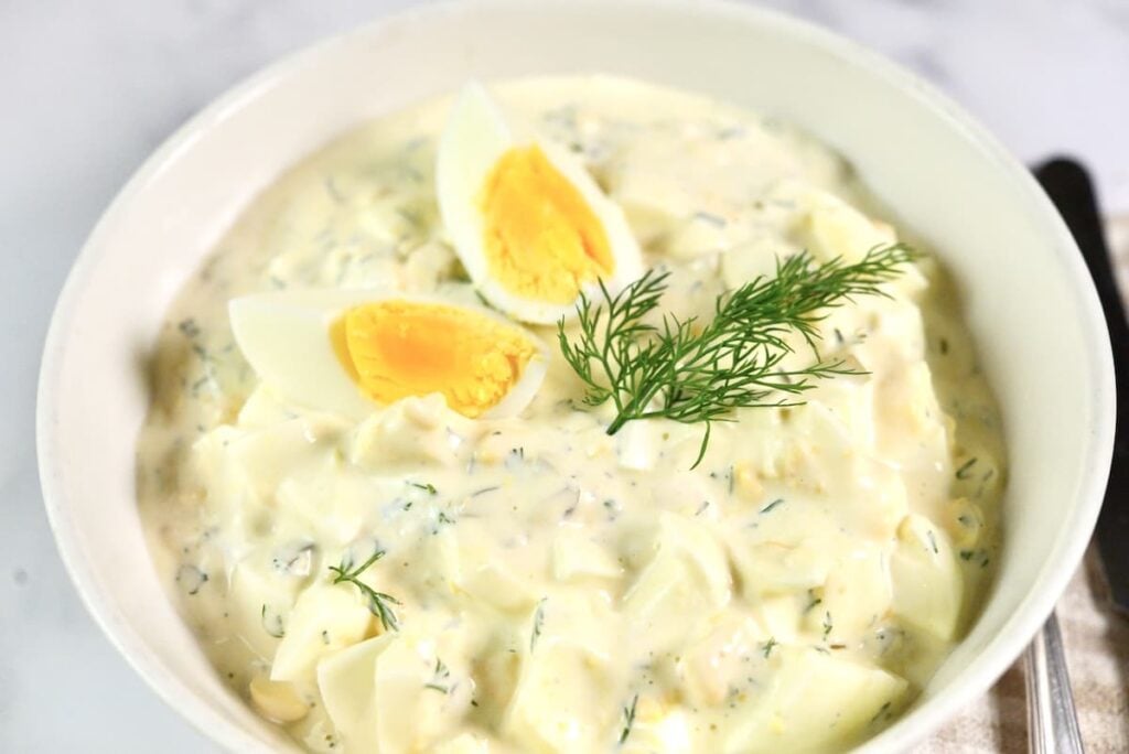 A bowl of creamy egg salad garnished with two quartered boiled eggs and fresh dill on top, served in a white dish on a light-colored surface.