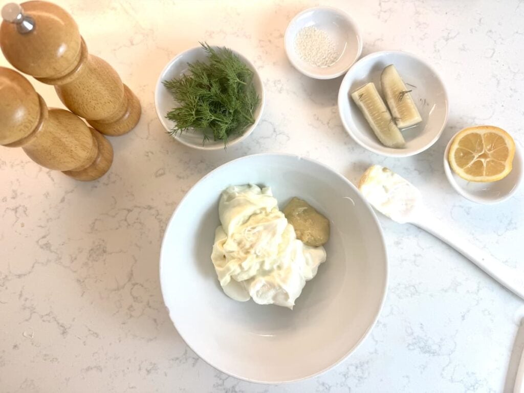 A white bowl with mayonnaise and mustard sits on a white countertop, surrounded by small bowls of fresh dill, sugar, pickles, half a lemon, and two wooden pepper and salt mills for eiersalat.