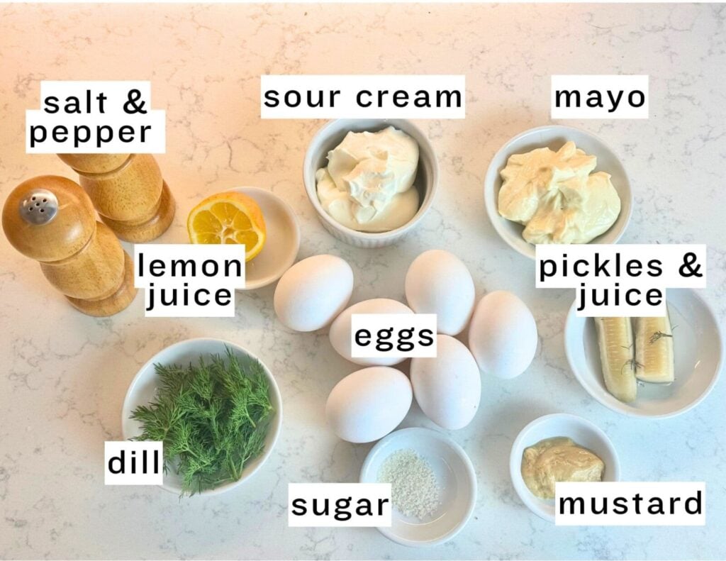 Ingredients for Eiersalat on a white countertop: salt and pepper shakers, a lemon wedge, fresh dill, sugar, six eggs, bowls of sour cream, mayo, pickles with juice, and mustard, each labeled.