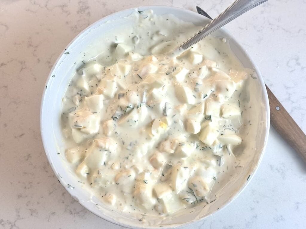 A white bowl filled with creamy egg salad containing chopped eggs, herbs, and mayonnaise, with a spoon resting in the bowl on a light-colored countertop.