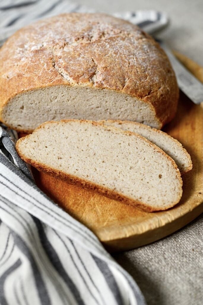 A round loaf of rustic bread with a golden crust sits on a wooden board. Two thick slices are cut and laid next to the loaf. A striped cloth is partially visible beside the bread.
