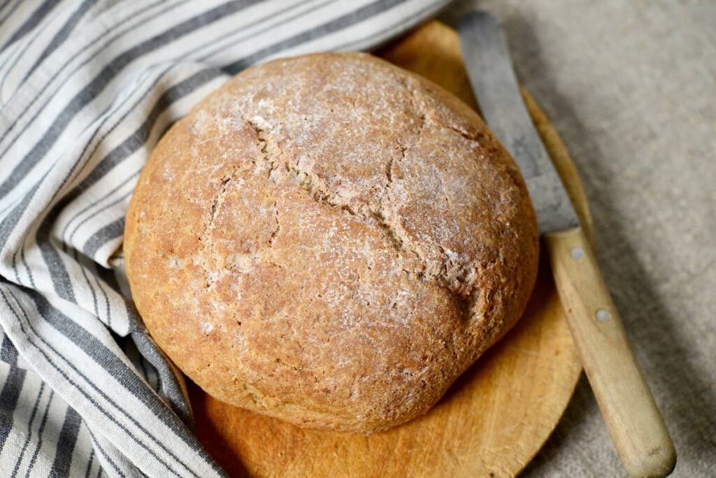 A round loaf of rustic bread dusted with flour sits on a wooden cutting board beside a serrated knife and a striped cloth.