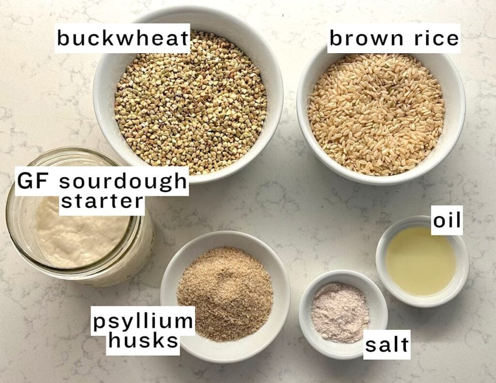 Six labeled bowls and jars on a white counter containing buckwheat, brown rice, gluten-free sourdough starter, oil, psyllium husks, and salt.