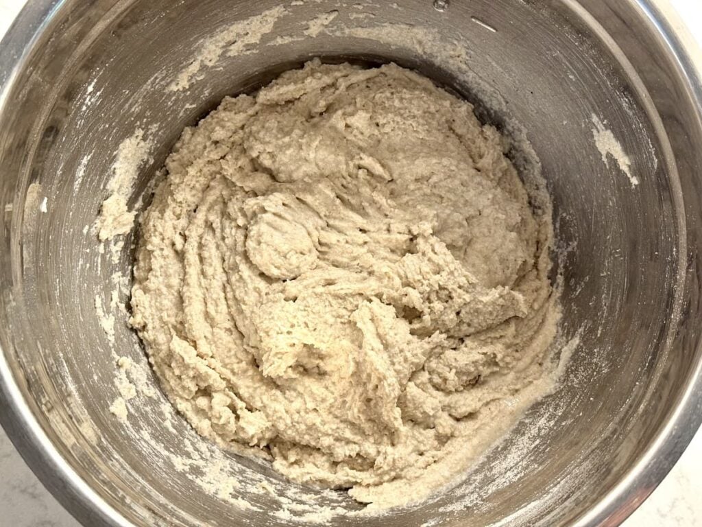 A metal mixing bowl filled with thick, beige bread dough that has been mixed and is ready for further preparation. The sides of the bowl have remnants of dough sticking to them.