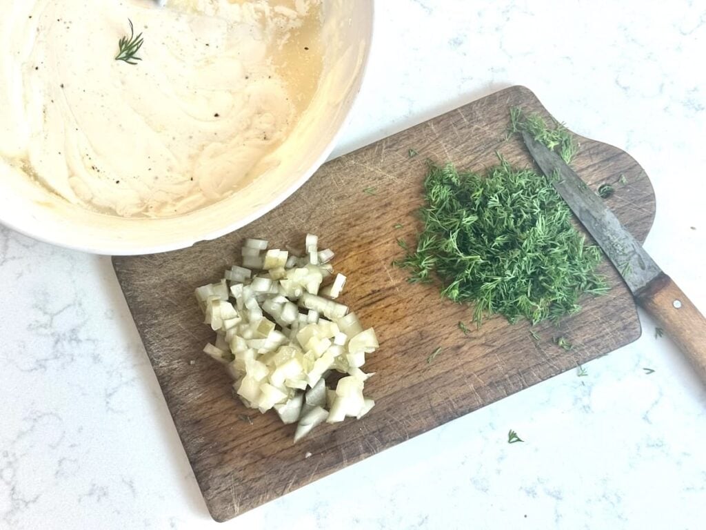 A wooden cutting board holds chopped pickles, chopped dill, and a knife. Beside it is a white bowl containing a creamy mixture with a sprig of dill on top, all on a light countertop.
