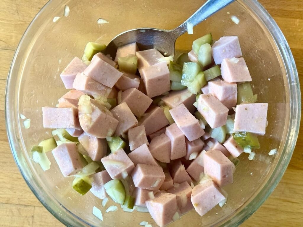 A glass bowl filled with a salad made of cubed pink luncheon meat, chopped pickles, and diced onions, with a metal spoon resting inside. The bowl is on a wooden surface.