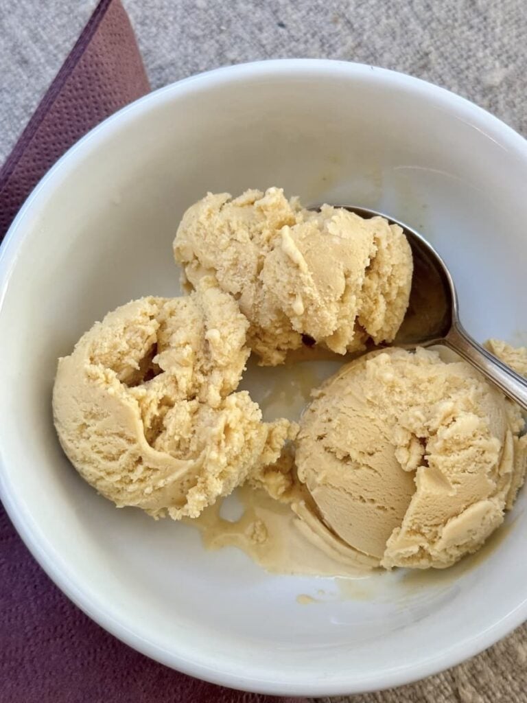 Three scoops of light brown ice cream in a white bowl with a metal spoon, placed on a textured surface next to a folded dark napkin.