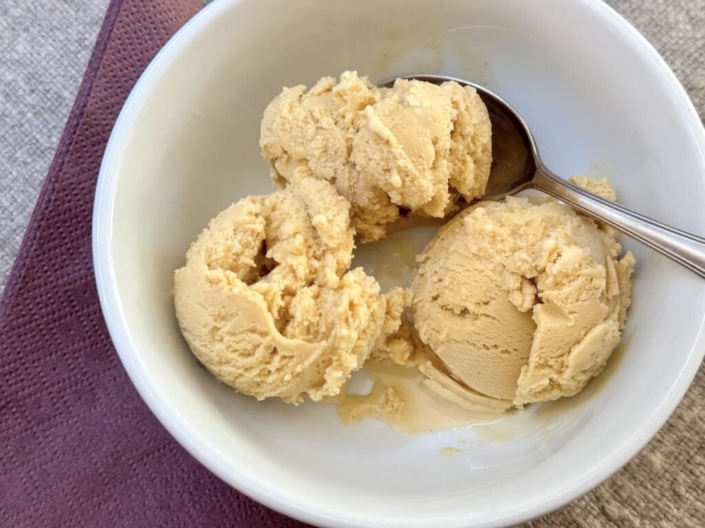 A white bowl with three scoops of light brown ice cream and a silver spoon, placed on a purple napkin on a textured surface.