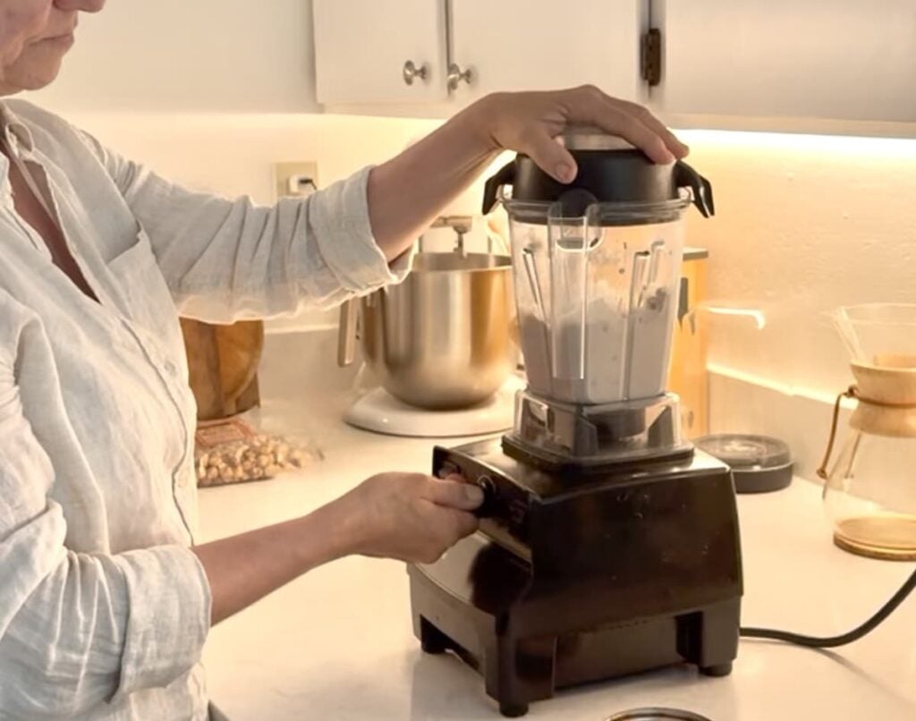 A person wearing a light-colored shirt is using a black countertop blender filled with a creamy mixture in a bright kitchen. Their hand is on the lid, and they are turning a dial to blend.