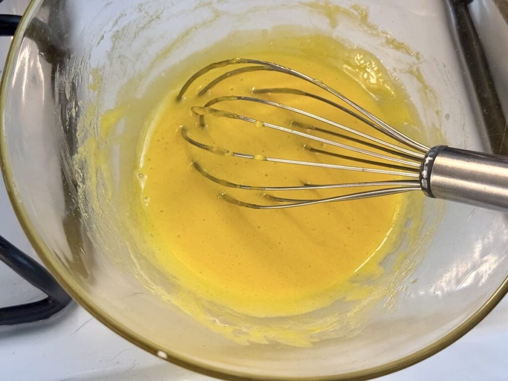 A metal whisk rests in a glass bowl filled with a smooth yellow batter, positioned on a white countertop near a stovetop burner.