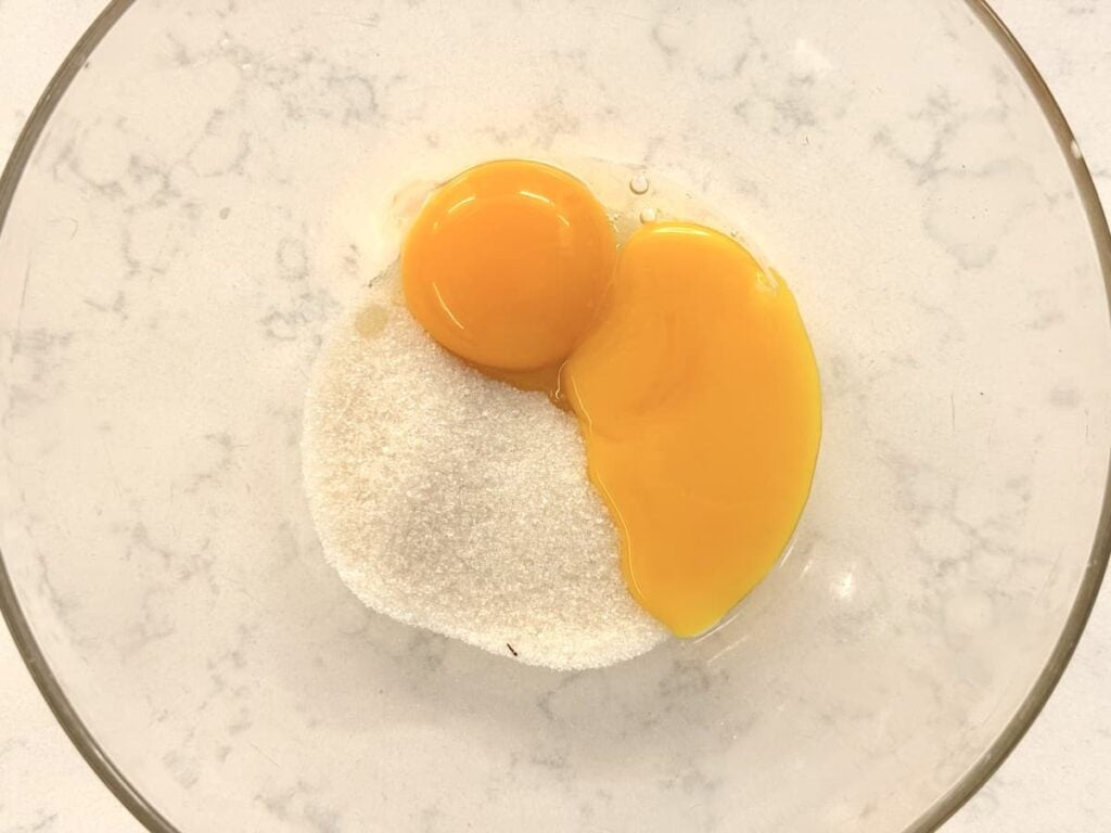 A glass bowl containing one whole raw egg, one raw egg yolk, and a mound of granulated white sugar on a white marble surface.
