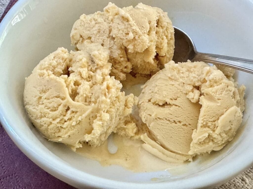 A white bowl containing three scoops of creamy, light brown ice cream with a metal spoon resting inside the bowl.