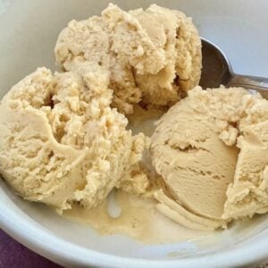 A white bowl containing three scoops of creamy, light brown ice cream with a metal spoon resting inside the bowl.