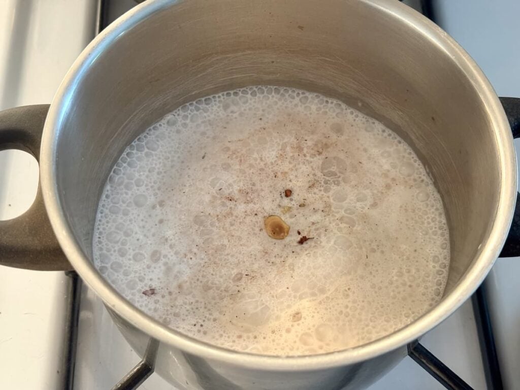 A stainless steel pot on a stove contains simmering milk with spices, producing bubbles and foam on the surface.