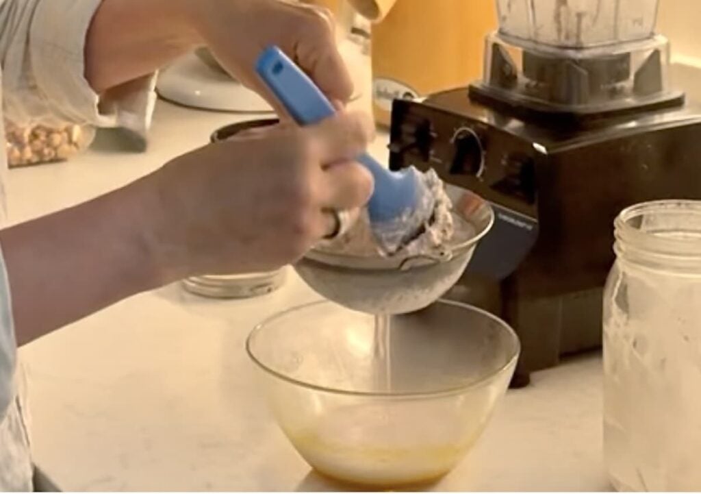 A person strains a blended mixture through a metal sieve into a glass bowl, using a blue spatula. A blender and glass jar are on the counter nearby.