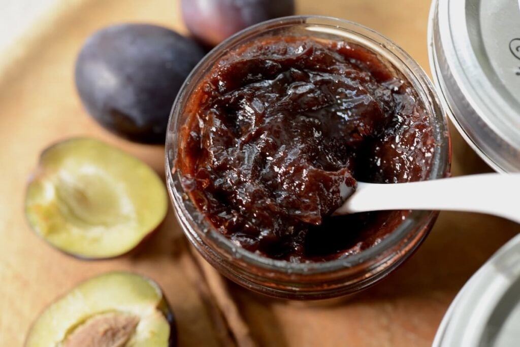 An open jar of dark German plum butter with a white spoon inside, surrounded by whole and halved plums on a wooden surface.