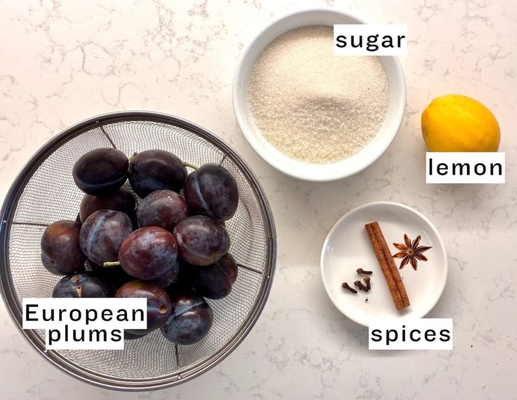 A bowl of European plums, a bowl of sugar, a whole lemon, and a small dish with cinnamon, cloves, and star anise—all labeled—are arranged on a white countertop, ready to be transformed into traditional German plum butter.