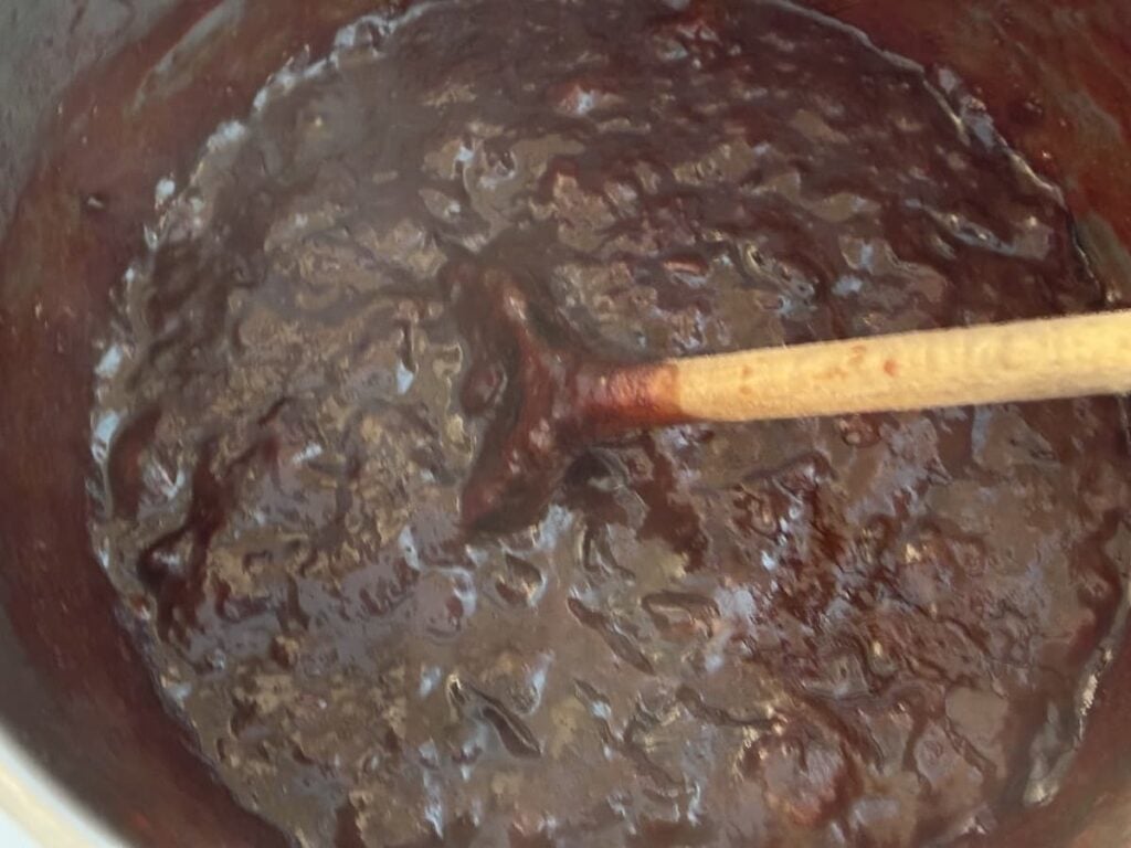 A wooden spoon stirs a thick, dark brown mixture in a pot, reminiscent of chocolate or fudge being cooked—or perhaps even German plum butter as it bubbles and deepens in color.