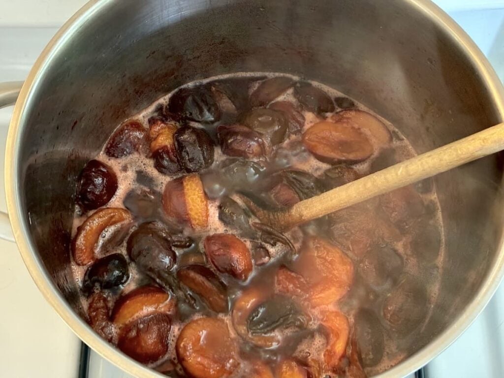 A pot of simmering fruit, including dark plums and peaches, being stirred with a wooden spoon on a stove. Steam rises from the bubbling mixture as it slowly transforms into a rich German plum butter.