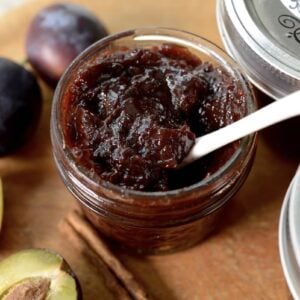 An open jar of dark plum jam, reminiscent of traditional German plum butter, with a white spoon inside, is surrounded by whole and halved plums, cinnamon sticks, and closed jars on a wooden surface.