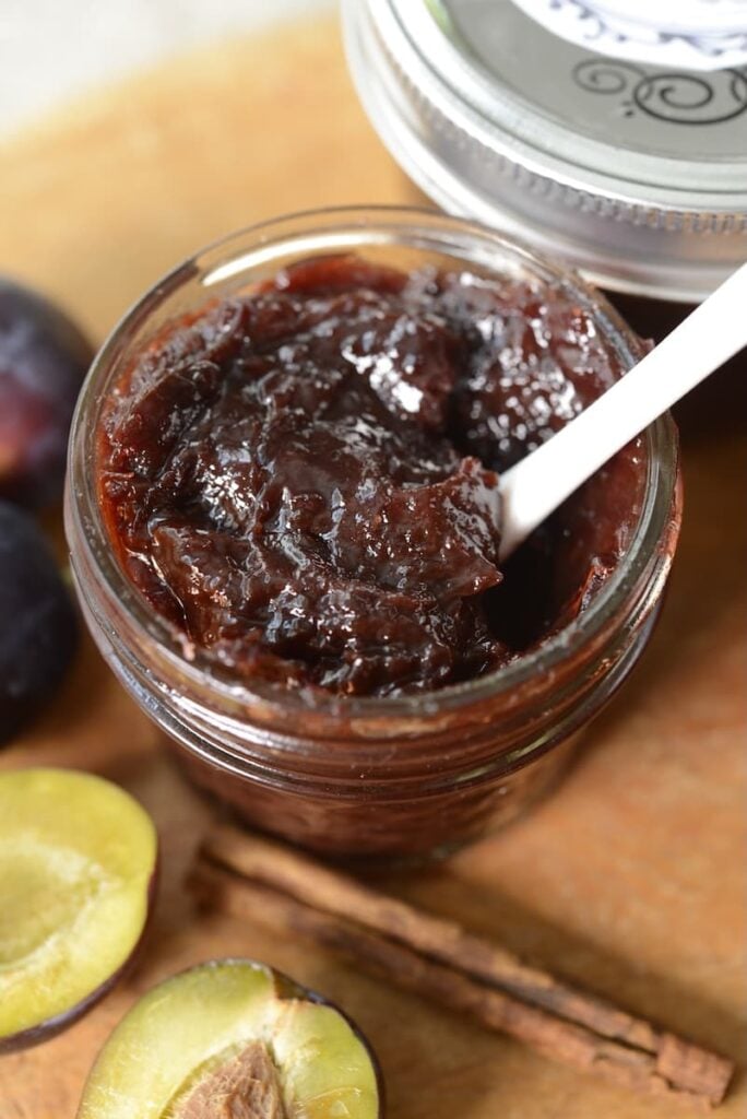 An open glass jar filled with thick, dark German plum butter, with a white spoon inside. Fresh plums, cinnamon sticks, and a closed jar lid are nearby on a wooden surface.