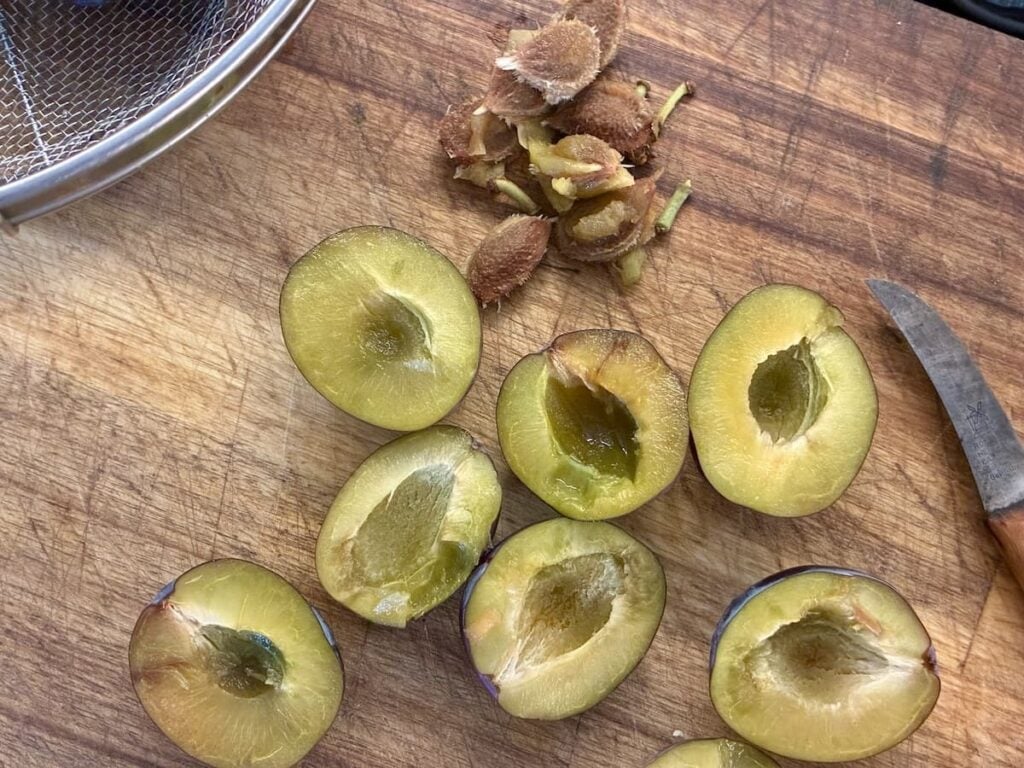 Halved plums with pits removed are on a wooden cutting board, ready for making traditional German plum butter. A small pile of pits, a paring knife, and part of a metal strainer are also visible.