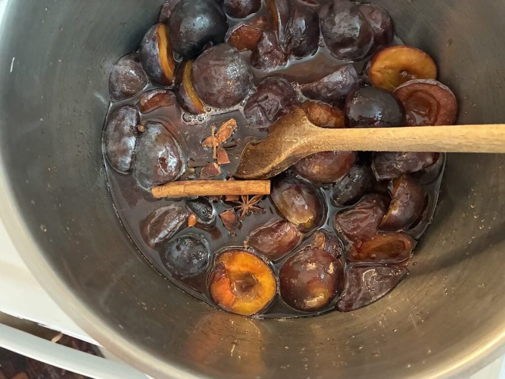 A pot of halved plums simmering in liquid with a wooden spoon, visible cinnamon stick, and star anise, evokes the traditional process of making German plum butter, likely being prepared for a rich jam or sauce.