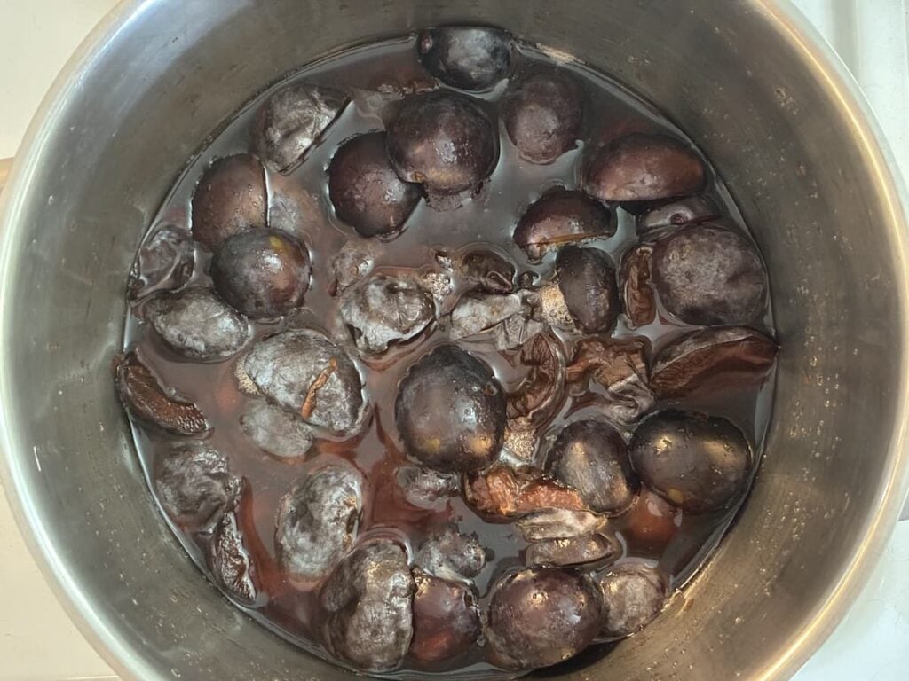 A pot filled with boiling dark purple plums, some whole, some split, simmering in their juices as they transform into rich German plum butter.