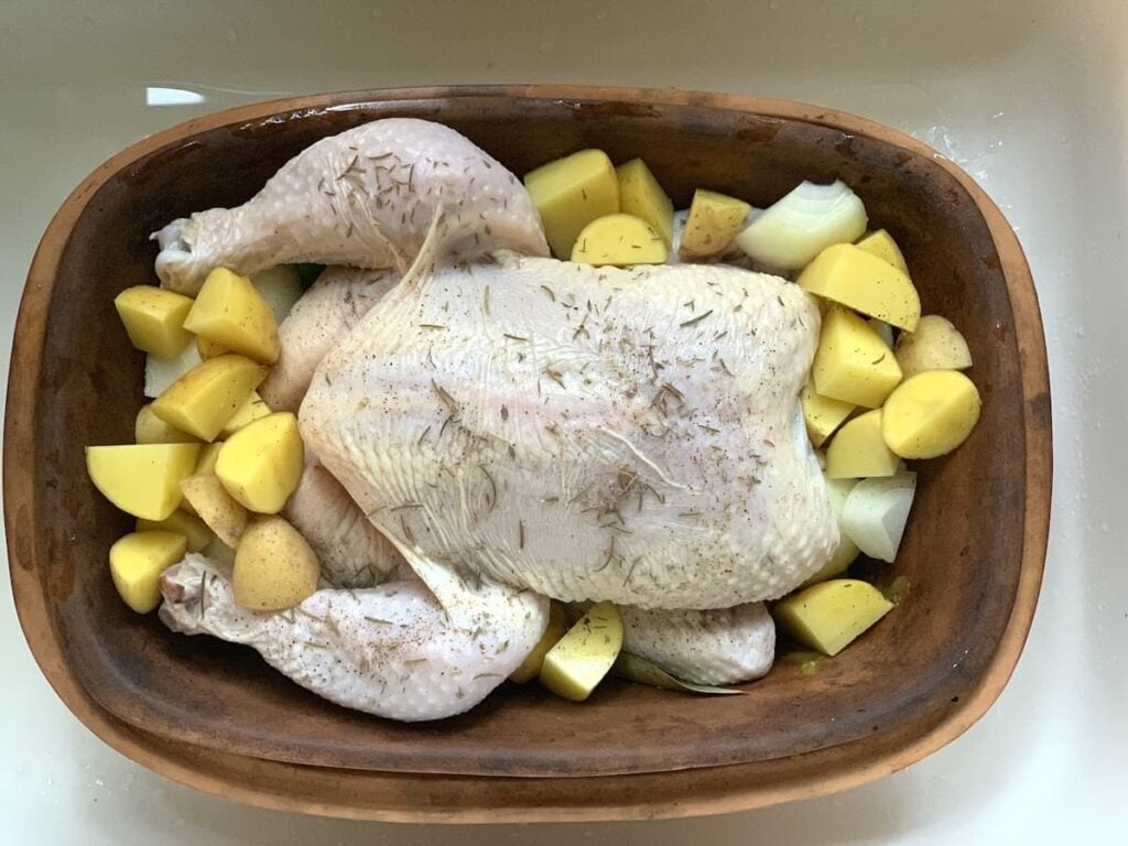 Raw whole chicken seasoned with herbs, surrounded by chopped yellow potatoes and onions, placed in a brown roasting dish ready for cooking.