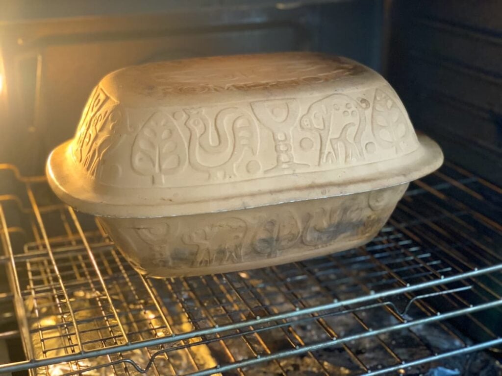 A decorated clay baking dish with a lid, featuring carved animal and tree designs, sits on a rack inside an oven.