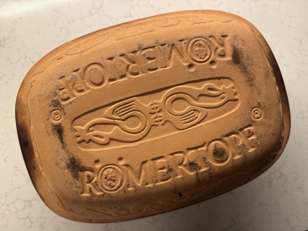 A terracotta Römer-topf baking dish with its brand name and decorative patterns engraved on the lid, placed on a light marble surface.