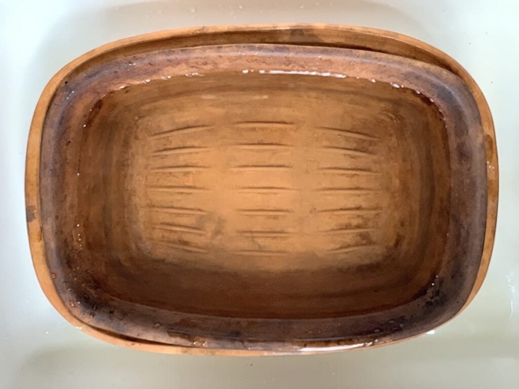 A rectangular, brown ceramic baking dish with visible scratches inside sits in a white sink, partially filled with water.