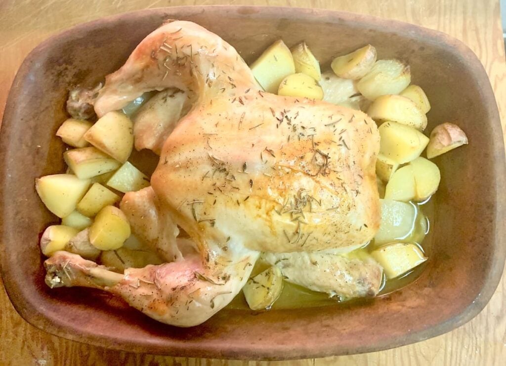 A whole roasted chicken with golden skin, surrounded by roasted potatoes and onions in a brown baking dish, all seasoned with herbs.