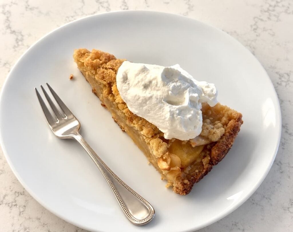 A slice of apple crumb cake with fruit crumble topping and whipped cream on a white plate with a fork.