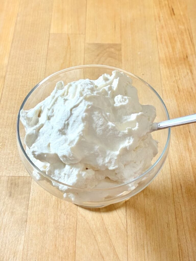 A glass bowl filled with whipped cream and a metal spoon on a wooden surface.