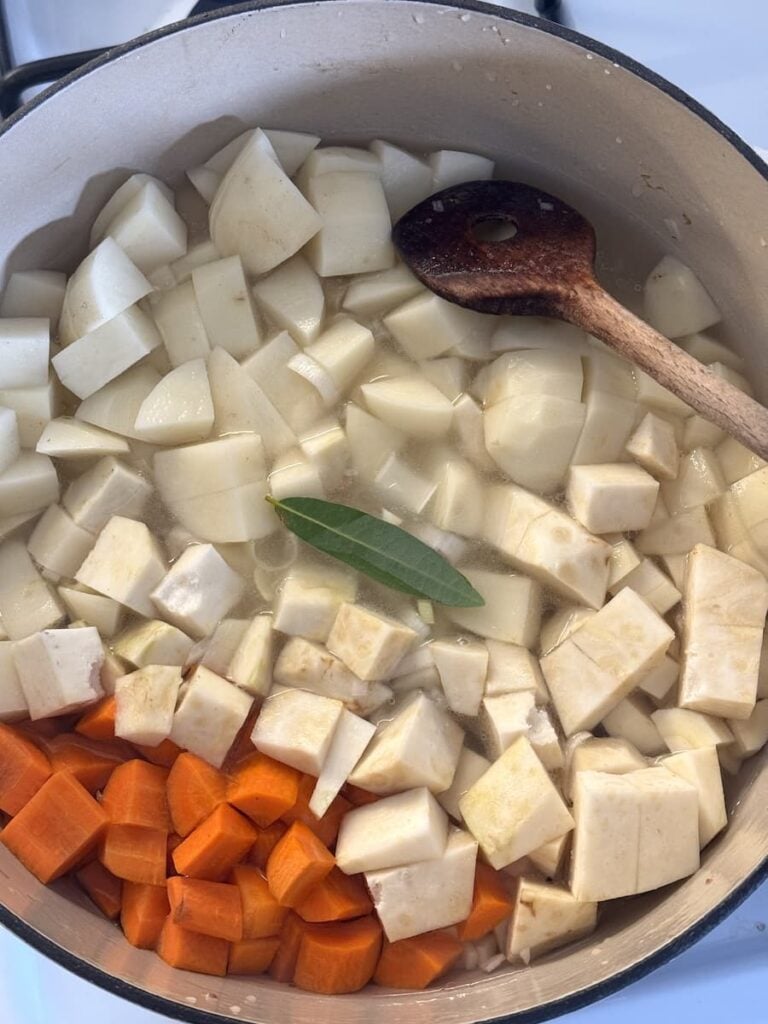 Chopped potatoes, parsnips, and carrots for a potato soup with a bay leaf in a pot of water, with a wooden spoon resting on the edge.