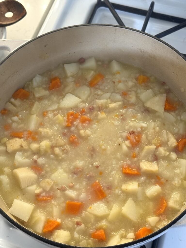 A thick potato soup with diced carrots and small bits of meat simmering in a large white pot.