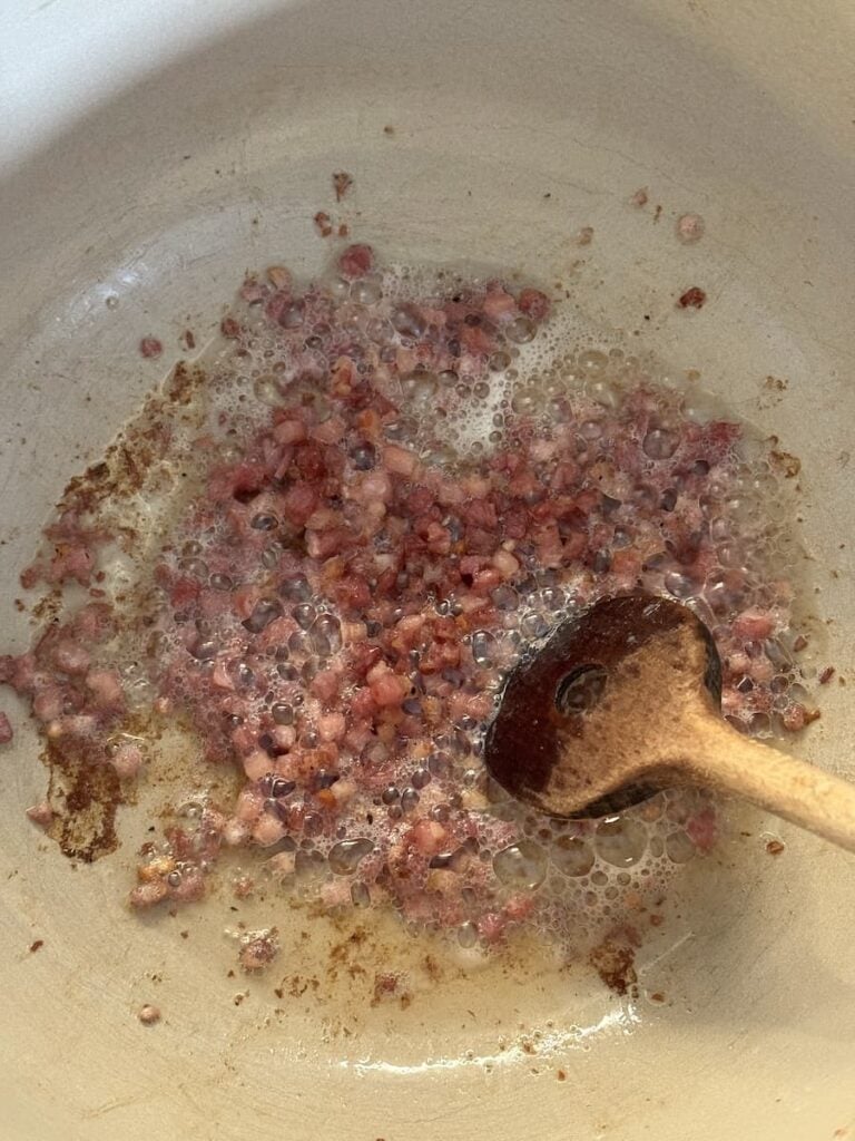 Diced bacon for potato soup sizzling in a skillet, being stirred with a wooden spoon.
