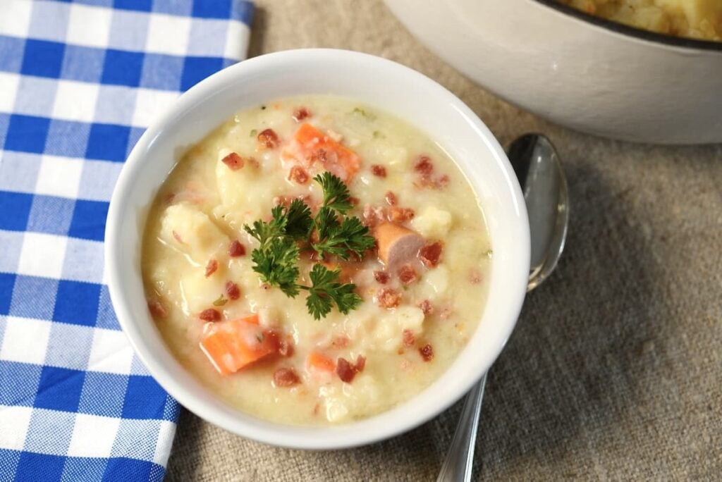 A bowl of creamy potato soup topped with bacon bits, sliced sausage, and parsley.
